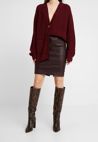 Femme portant un cardigan en tricot oversize bordeaux, une jupe en cuir marron foncé et des bottes hautes en cuir imprimé serpent marron se tenant sur un sol blanc.