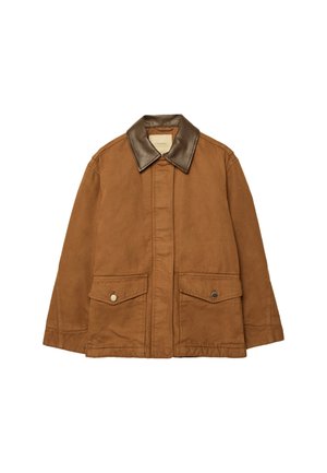 Personne portant une veste marron, une chemise rayée, un short en jean et des bottes noires montantes, debout sur un sol carrelé contre un mur gris.