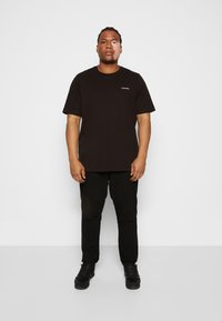 Sort bomulds t-shirt med korte ærmer og rund udskæring, prydet med et lille hvidt logo på brystet, sammen med sorte bukser og sko.