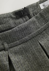 Pelēka zivju kaula raksta audums ar pogu un rāvējslēdzēju aizdari, ar piesietu jostas detaļu un krokojumiem pie jostasvietas.