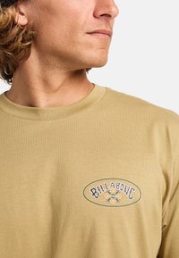Beige overhemd met lange mouwen en een ronde hals, voorzien van een ronde Billabong-logo geborduurd in blauw en geel op het borstgebied.
