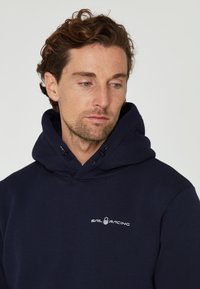 Sudadera azul marino hecha de una tela suave, con capucha ajustable y un logo blanco de "Sail Racing" en el pecho.