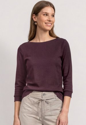 Femme souriante portant un haut à manches longues violet foncé et un pantalon beige clair avec une taille à cordon sur un fond uni.
