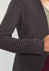 Närbild av en person som bär en svart jacka med ett litet geometriskt rosa och vitt mönster över en olivgrön topp.