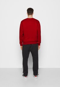 Sudadera roja con cuello redondo, pantalones de chándal negros con logotipos laterales y zapatillas grises, vistos desde la parte posterior sobre un fondo liso.