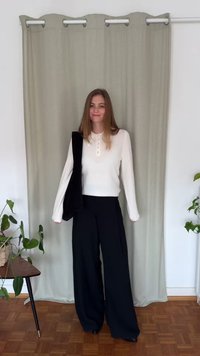 Biała koszulka polo z długim rękawem i guzikami, zestawiona z luźnymi czarnymi spodniami. Widoczna czarna torebka na ramię i rośliny doniczkowe.