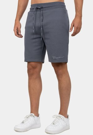 Man draagt donkergrijze shorts met trekkoord en witte sneakers, staand tegen een effen lichte achtergrond.