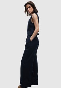Vrouw in een marineblauw gestreept mouwloos vest en bijpassende wijde broek, staand in zijprofil met één hand in de zak tegen een effen achtergrond.
