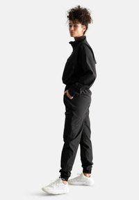Tuta sportiva nera con colletto zip, polsini e vita elasticizzati. Abbinata a scarpe da corsa bianche, che mettono in evidenza una silhouette aerodinamica.