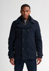 Cappotto di lana blu navy con chiusura a doppio petto, ampi rever e polsini regolabili. Presenta uno strato interno con zip.