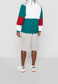 Kolorowa kurtka przeciwwiatrowa z turkusowymi, białymi, czerwonymi i beżowymi fragmentami; elastyczne mankiety; zestawiona z beżowymi szortami typu cargo i białymi trampkami.