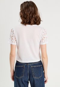 Haut blanc à manches courtes avec des accents en dentelle aux épaules ; tissu doux ; longueur cropped ; associé à un jean en denim bleu, montrant la vue de dos.