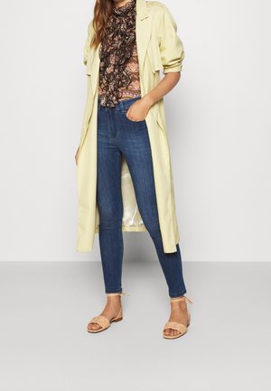 Lichtgele trenchcoat over een gepatterne top, gecombineerd met donkerblauwe jeans en beige sandalen. De jas heeft opgerolde mouwen en een relaxte pasvorm.