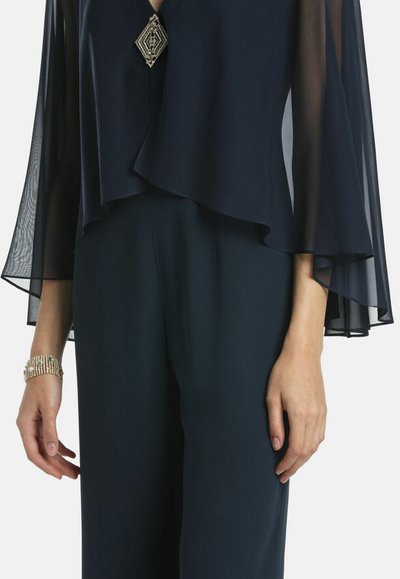 Marineblaues Overlay-Oberteil mit durchsichtigen langen Ärmeln und einem geometrischen silbernen Akzent am Ausschnitt, kombiniert mit dunkelblauen, weit geschnittenen Hosen.