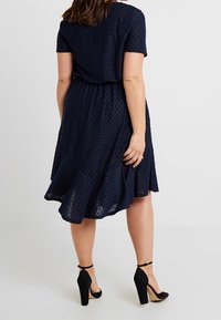 Mörkblå klänning med korta ärmar, strukturerat prickmönster, åtsittande midja och utsvängd nederkant, kombinerad med svarta högklackade skor.