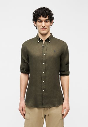 Young man with curly hair wearing an olive green buttoned shirt with rolled-up sleeves and beige cargo pants, standing against a plain background.