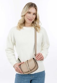 Borsa a tracolla in pelle beige con una patella curva, hardware circolare dorato e una sottile tracolla regolabile, abbinata a un maglione bianco e jeans blu.