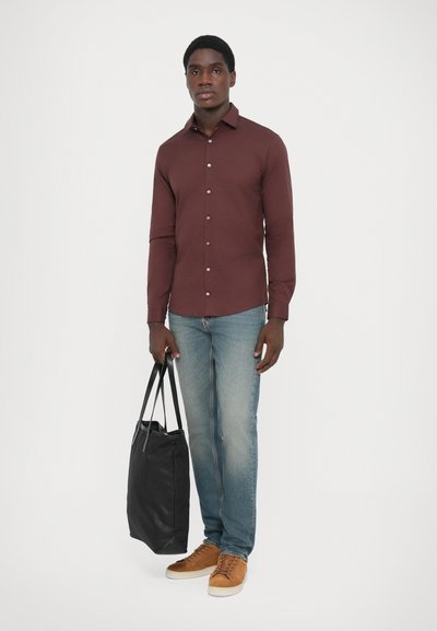 Camisa de manga larga burdeos con botones, jeans azules y zapatillas marrones. Sosteniendo una bolsa de tote negra, de pie contra un fondo blanco liso.