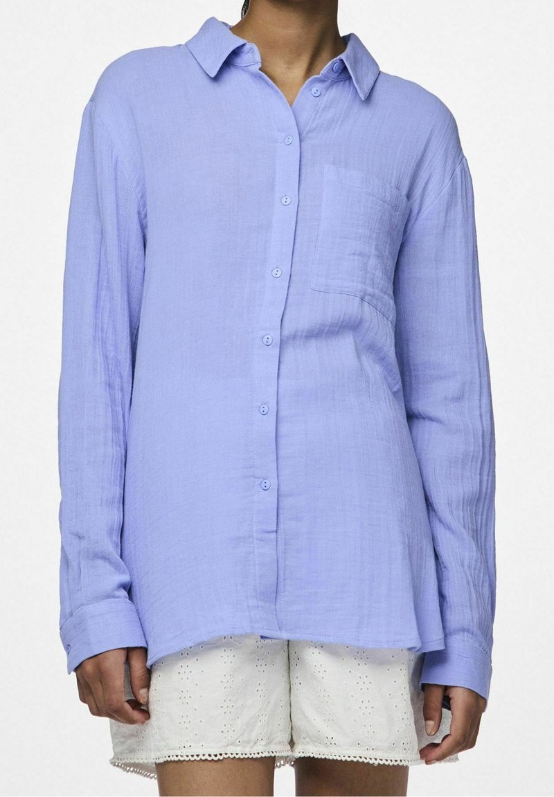 Personne portant une chemise ample à boutons à manches longues de couleur violet clair et un short en dentelle ajourée blanc, debout devant un fond uni.
