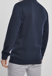 Felpa blu navy in tessuto morbido, con maniche lunghe, scollo e orlo a coste. Mostrata da dietro, abbinata a pantaloni della tuta grigi.