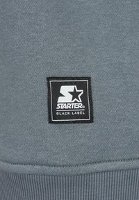 Tessuto felpato grigio con superficie strutturata, caratterizzato da una toppa quadrata con logo in bianco e nero con la scritta "STARTER BLACK LABEL".