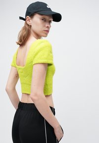 Pulover scurt de culoare verde lime, dintr-un material texturat, cu mâneci scurte și spate pătrat, asortat cu pantaloni sport negri, cu o dâra laterală.