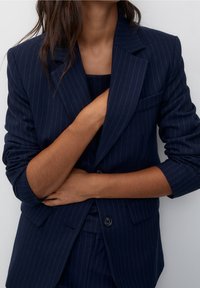 Femme portant un costume bleu marine à fines rayures avec les manches du blazer retroussées, les bras croisés et la main posée sur la poitrine.