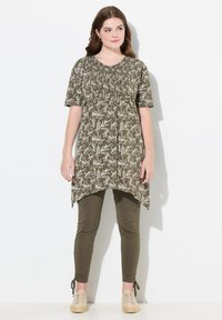 Tunique vert olive avec imprimé floral blanc, avec des manches courtes et un corsage élastiqué ; assortie à des leggings vert olive et des chaussures beiges.