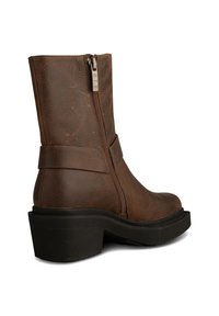 Bota de tobillo de cuero marrón con cierre de cremallera, una correa ancha alrededor del tobillo y un tacón de goma negra gruesa con surcos texturizados.