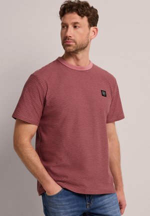 Mann mit kurzen lockigen braunen Haaren, der ein rot-schwarz gestreiftes kurzärmeliges T-Shirt und blaue Jeans trägt, steht mit der rechten Hand in der Tasche.