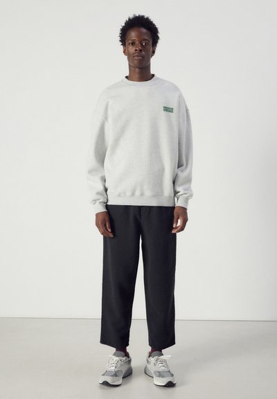 Personne debout portant un sweat-shirt gris clair avec le texte "American Vintage", un pantalon court noir et des baskets blanches et grises.