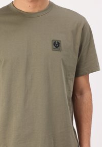 Ελιά πράσινη κοντομάνικη μπλούζα φτιαγμένη από μαλακό βαμβάκι, με ένα μαύρο ορθογώνιο λογότυπο στην αριστερή πλευρά του στήθους.