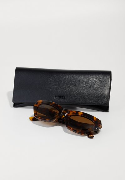Tortoiseshell sunglasses with dark lenses and angular frame, placed next to a black leather case, featuring a smooth texture and folded design.