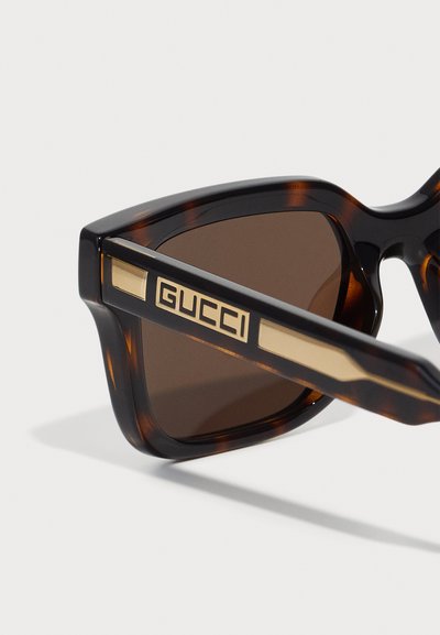 Gucci solbriller med skilpaddeskallramme, brune linser og dristig gullakcent ved tempelet som viser merkenavnet.