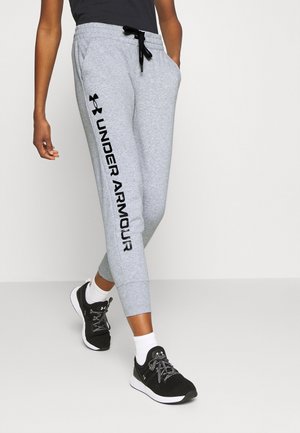 Osoba nosząca szare spodnie dresowe Under Armour z logo na nodze, czarne trampki, białe skarpetki i czarny top, idąca po jasnej podłodze.