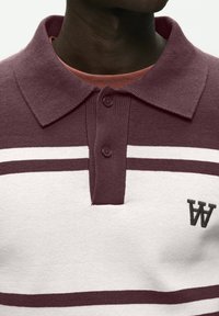 Nærbillede af en person, der bærer en bordeaux og hvid stribet polo-trøje med to knapper og et lille broderet "WA"-logo.