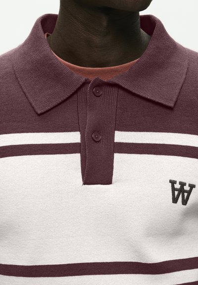 Gros plan sur une personne portant une chemise en tricot à col, rayée bordeaux et blanche, avec deux boutons et un petit logo brodé "WA".