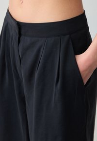 Pantalons plissés noirs avec une texture douce, dotés d'une taille et de poches latérales, mettant en avant une coupe décontractée et un design épuré.