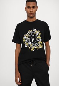 Black cotton crew-neck t-shirt featuring a colorful circular logo design with gold accents and intricate patterns. Paired with black trousers.