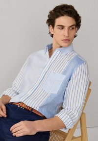 Giovane uomo seduto su una sedia di legno, indossa una camicia a righe verticali blu e bianche con bottoni e pantaloni blu navy con cintura marrone, guarda verso destra.