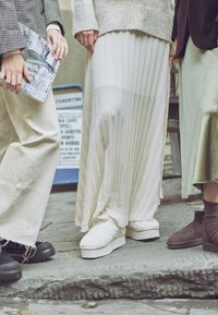 Tři lidé stojící na kamenném schodu, oblečení v různých sukních a texturované obuvi, z nichž jeden drží časopis s názvem "Sensi".