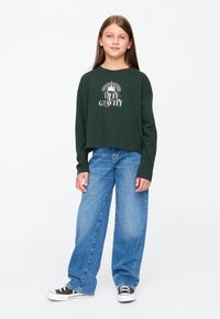 Zöld, crop fazonú, hosszú ujjú póló "DEFY GRAVITY" felirattal, laza kék farmernadrággal és fekete, fehér részletekkel díszített tornacipővel párosítva.