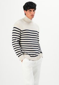 Uomo che indossa un maglione a collo alto a righe bianche e blu navy e pantaloni bianchi, in piedi con le mani in tasca contro uno sfondo semplice.