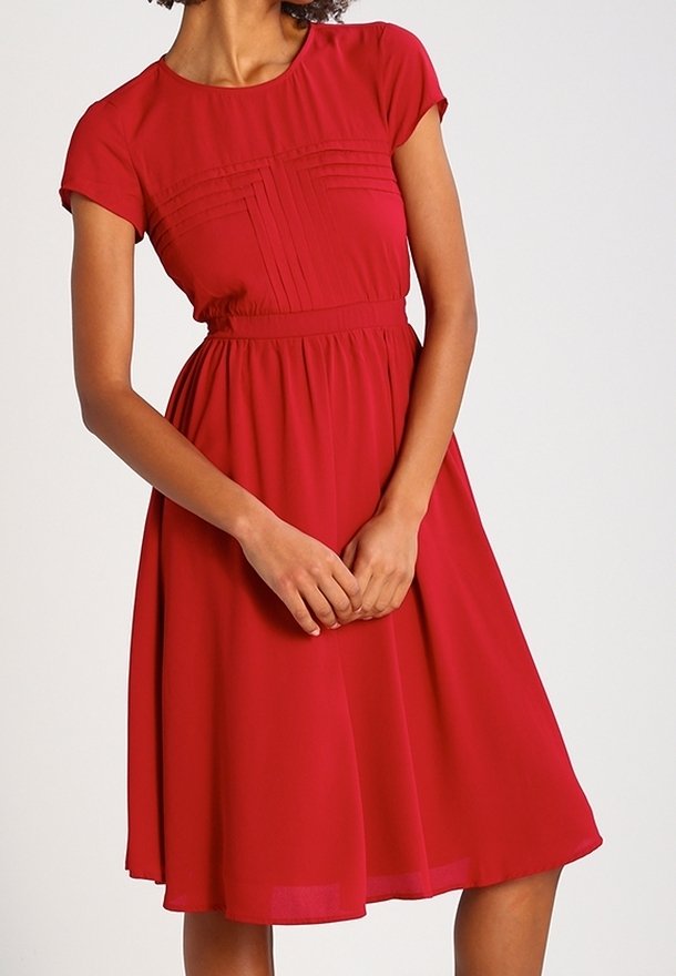 Femme portant une robe rouge au genou, manches courtes, taille cintrée et détails plissés sur la poitrine, debout, mains jointes.