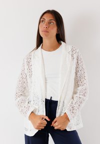 Blazer bianco in pizzo con maniche interamente in pizzo, rever a punte e design con apertura anteriore. Indossato sopra una t-shirt bianca e pantaloni scuri.