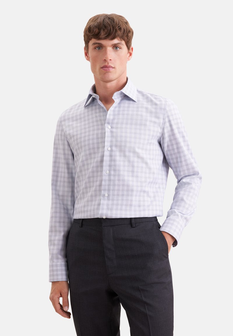 Jeune homme aux cheveux bruns courts portant une chemise à carreaux gris clair à manches longues et un pantalon gris foncé, debout, une main dans la poche.