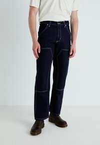 Dark denim jeans with contrast stitching, front pockets, and a straight-leg design. Paired with brown boots and a light-colored top.