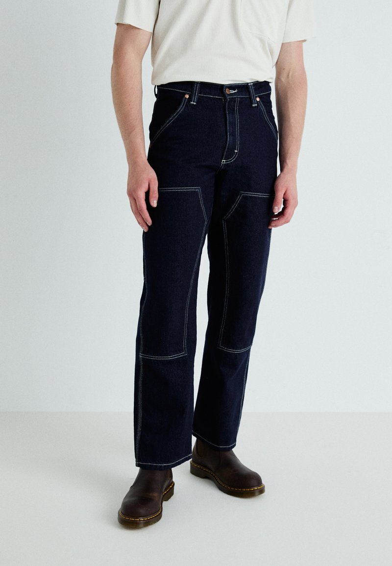 Dark denim jeans with contrast stitching, front pockets, and a straight-leg design. Paired with brown boots and a light-colored top.