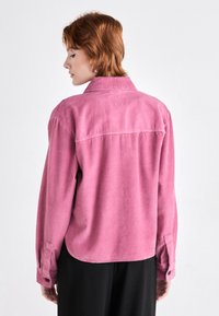 Růžová kordurová košile s zadním sedlem, dlouhými rukávy a manžetami na knoflíky. Obsahuje vertikální ribbing a zakřivený lem.