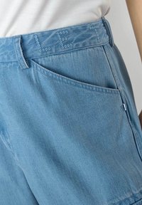 Shorts in denim azzurro chiaro con vita media. Presentano borchie decorative blu lungo la cintura e una tasca laterale.